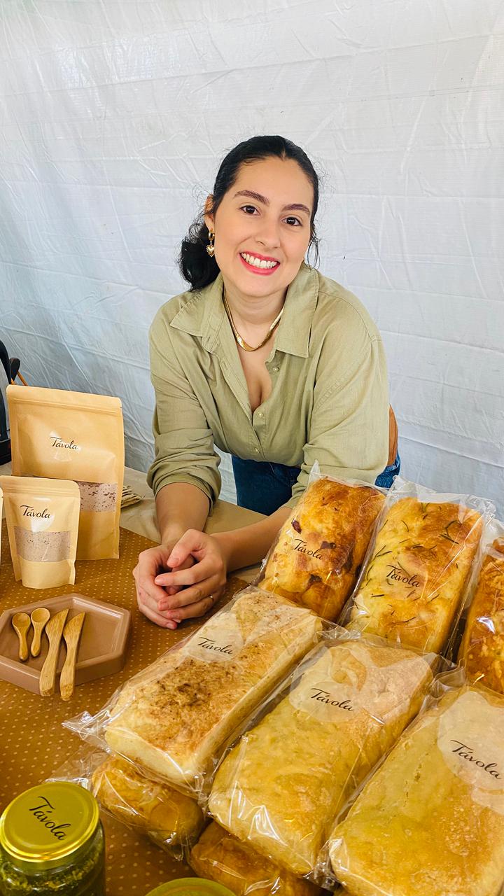Gabriella Tanico sorrindo atrás da mesa da Tavola, cercada pelos pães e produtos da marca.